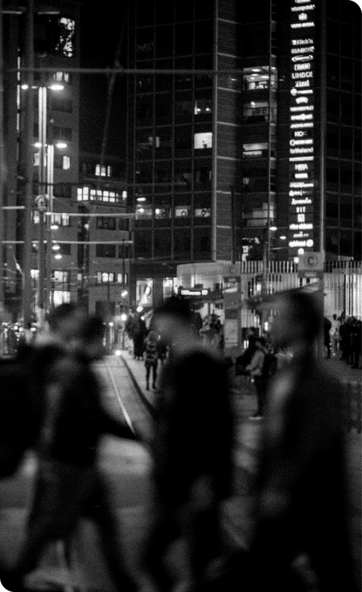 Svart/hvitt-bilde av et travelt bybilde kveldstid. Folk passerer i forgrunnen av bildet og i bakgrunnen, synes trikkeskinner, høye bygninger, flere folk og lysskilt.