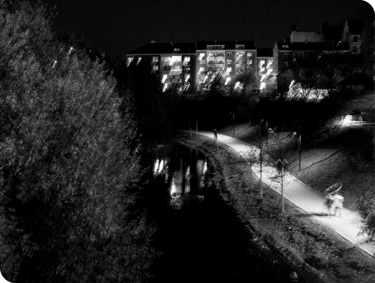 Svart/hvitt-bilde av en turvei som går langs en svingete elv. Det er en mørk kveld, og det går et par på turstien, og bygninger kan ses i bakgrunnen.