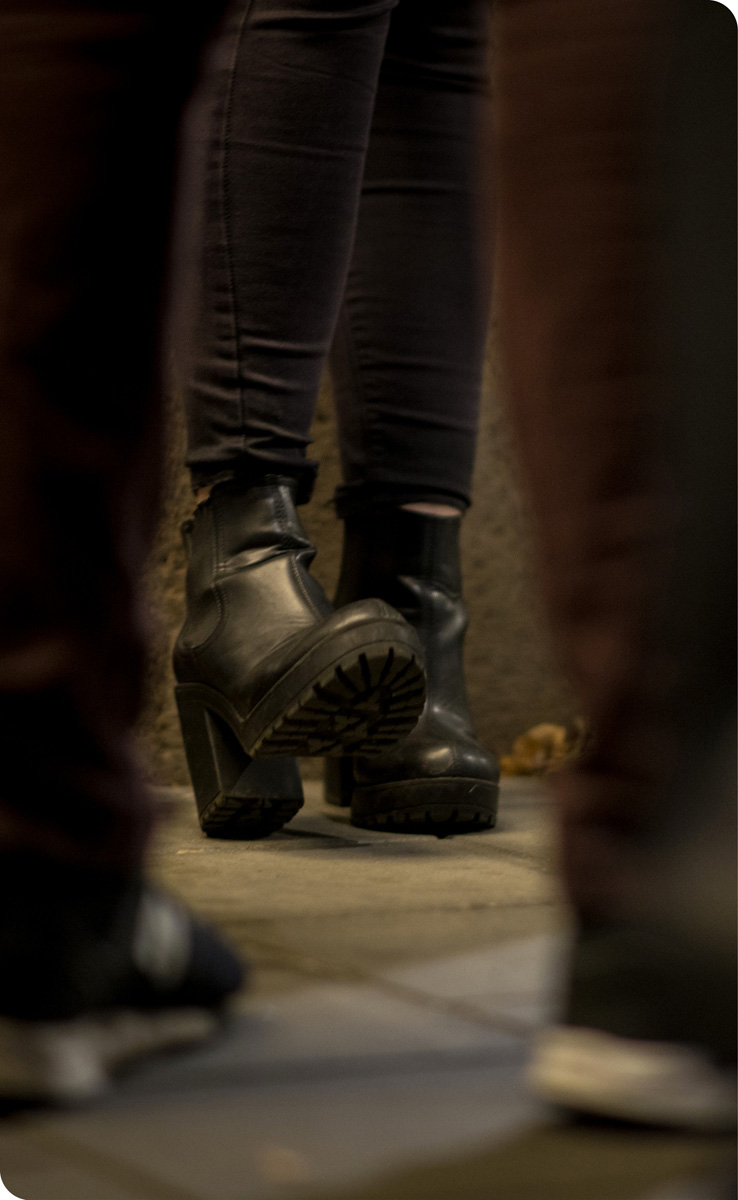 Bilde av ben med høyhælte skinnsko og trange bukser som står inntil en vegg. Det står en mann foran henne – og bilde er tatt mellom bena hans. Motivet antyder at det er en situasjon med en prostituert og en kunde eller hallik.