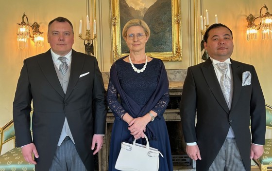 Bilde av tre ambassadører stående inne. Mennene i sjakett og kvinnen i kjole.