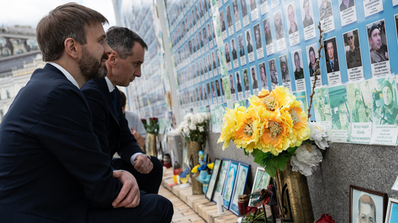 Jan Christian Vestre kneels in front of a wall with pictures of fallen Ukrainian soldiers. The Ukrainian Minister of Health also kneels beside Vestre.