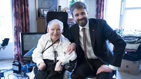 Til venstre sit Jostein i ein elektrisk rullestol, kledd i ei kvit skjorte og mørke bukser. Han har eit kommunikasjonsverktøy festa til rullestolen. Til høgre sit helse- og omsorgsminister Jan Christian Vestre, kledd i ein mørk dress og slips. Bakgrunnen viser eit romsleg rom med eit vindauge som slepp inn dagslys, og det er synlege møblar og personlege gjenstandar rundt i rommet.