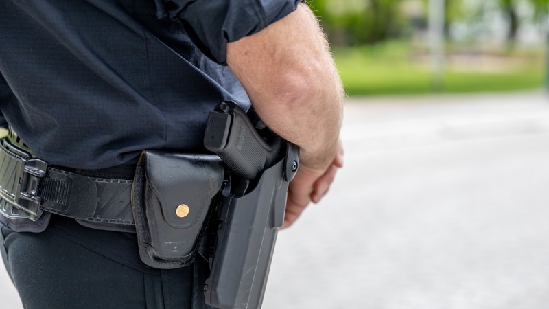 Nærbilde av midtpartiet til en person kledt i mørkeblå uniform. I beltet har vedkommende en pistol.