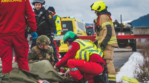 Fem personer fra med militær- og nødetatsuniformer behandler pasient på bakken. I bakgrunnen ser vi en ambulanse og et militært kjøretøy. Det er vinter.