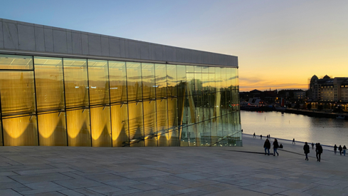 Vi ser Operahuset og en del av taket ned mot Oslofjorden en sommerkveld. Det er solnedgang og fin himmel i blått og guloransje. Samme farger sees også i vinduene på fasaden.