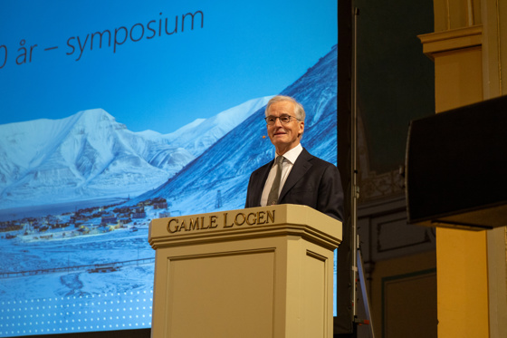 Statsminister Støre står bak en talerstol med påskriften Gamle Logen. Støre i dress og hvit skjorte med grått slips. Et stort lerret i bakgrunnen, med bilde fra Longyearbyen.